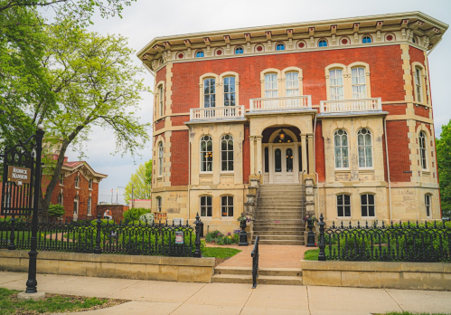 Historic red brick building with ornate architecture, large steps, and decorative iron fence, surrounded by greenery.