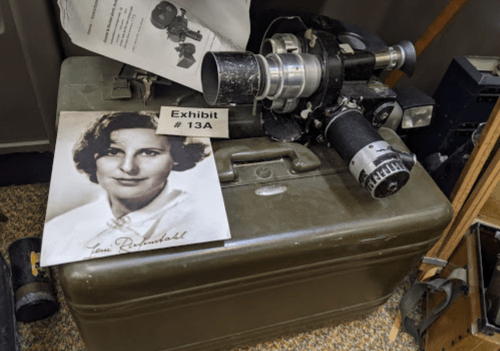 A vintage camera on a green case next to a black-and-white photo of a woman, labeled "Exhibit # 13A."
