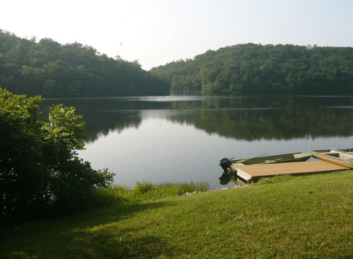 Serene lake view with calm water reflecting green hills, a grassy shore, and a small dock with boats.