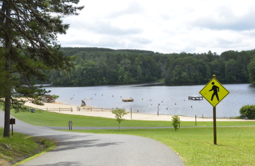 A peaceful lakeside scene with a sandy beach, boats, and a pedestrian crossing sign along a winding path.