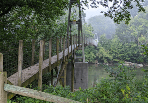 A wooden suspension bridge surrounded by lush greenery and mist, spanning over a calm river.