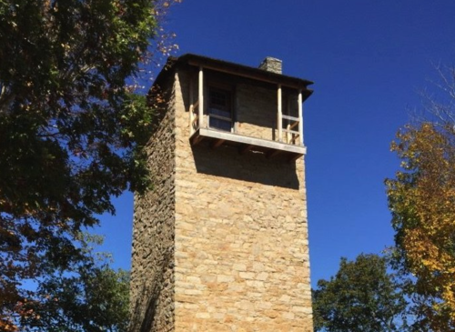 A tall stone tower with a wooden balcony, set against a clear blue sky and surrounded by trees.