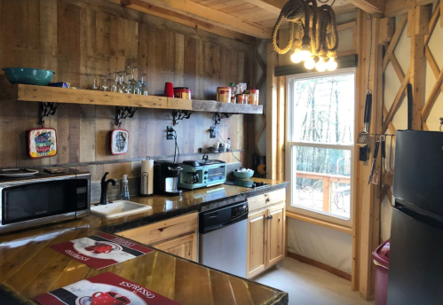 Cozy kitchen with wooden shelves, appliances, and a large window overlooking trees. Warm, rustic decor.