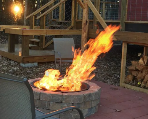 A vibrant fire burns in a stone fire pit, with a wooden structure and stacked logs in the background.