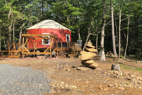 A red yurt surrounded by trees, with a stacked rock sculpture in the foreground and gravel path leading up to it.