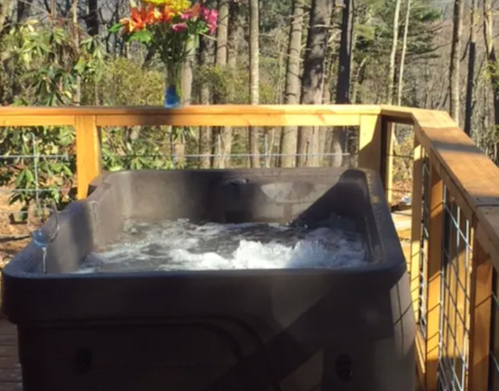 A hot tub on a wooden deck surrounded by trees, with a vase of flowers nearby.