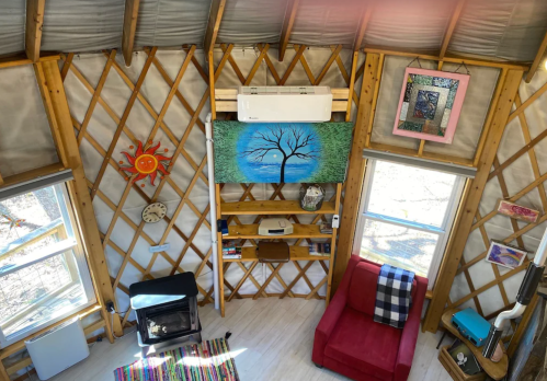 Interior of a cozy yurt featuring a red chair, artwork, a wood stove, and a tree painting on the wall.
