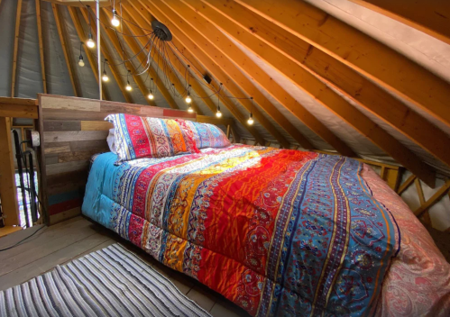 Cozy attic bedroom with a colorful patterned quilt, wooden beams, and warm string lights overhead.
