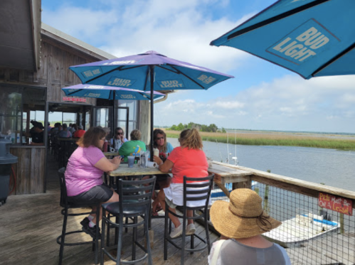 A group of people sitting at an outdoor bar by the water, enjoying drinks under blue umbrellas on a sunny day.