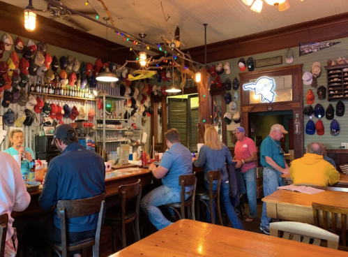 A cozy restaurant interior with patrons at the bar, surrounded by a wall of colorful hats and festive decorations.