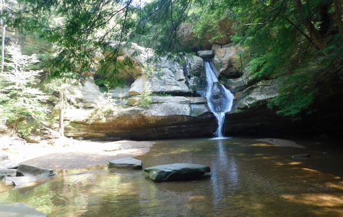 A serene waterfall cascades over rocks into a calm pool, surrounded by lush green trees and natural stone formations.