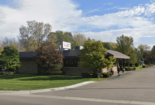 A stone building with a brown roof, surrounded by trees and grass, on a quiet street under a partly cloudy sky.
