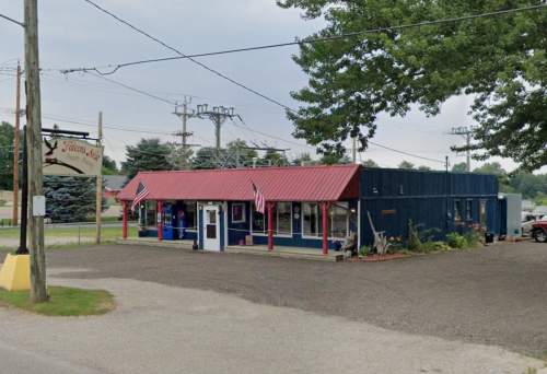 A small blue building with a red roof, featuring flags, surrounded by gravel and power lines.