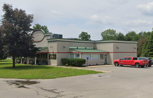 A single-story building with a green roof and red accents, surrounded by grass and trees, with a red truck parked outside.