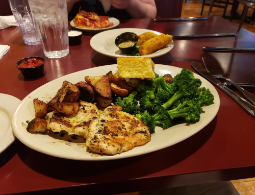 A plate of grilled fish with seasoned potatoes, broccoli, and cornbread, served with dipping sauce on a restaurant table.