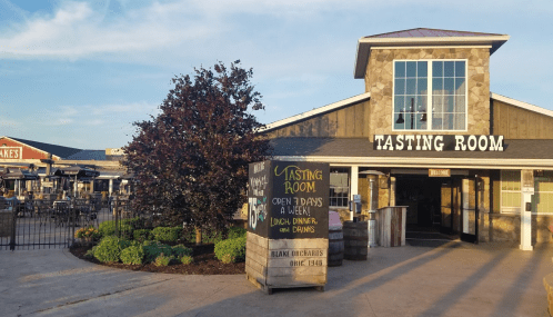 Exterior of a tasting room with a stone facade, featuring a sign that says "Open 7 days a week" and outdoor seating.