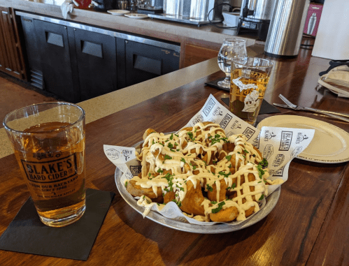 A plate of loaded fries drizzled with sauce, alongside a glass of cider and a beer at a bar.