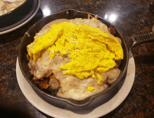 A skillet dish featuring a layer of yellow scrambled eggs over sausage, gravy, and potatoes.