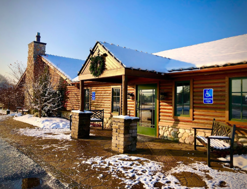 A log cabin-style building with snow on the roof, featuring a wreath and accessible entrance signs.