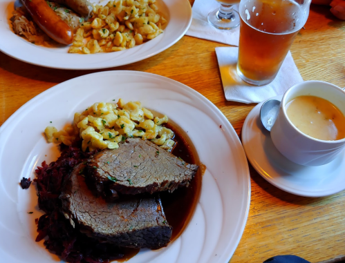 A plate with sliced beef brisket, red cabbage, and noodles, accompanied by a cup of gravy and a glass of beer.