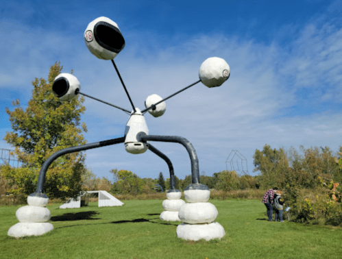 A large, abstract sculpture with spherical elements on long arms, set in a grassy area with trees and a blue sky.