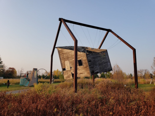 A tilted wooden house suspended by metal beams in a grassy area, with people walking nearby under a clear sky.