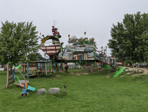 Colorful playground structure resembling a ship, surrounded by green grass and trees on a cloudy day.