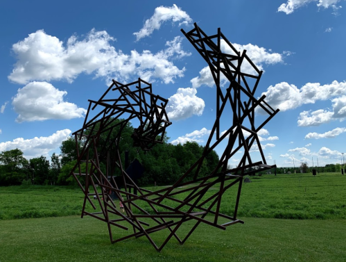 A large, abstract metal sculpture spirals upward against a blue sky with fluffy clouds, set in a green field.