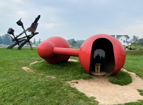 A large red sculpture resembling a dumbbell, with a small entrance, sits on green grass near another abstract sculpture.