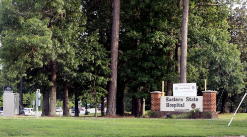 Sign for Eastern State Hospital surrounded by trees, with a bike rack and parking area visible in the background.