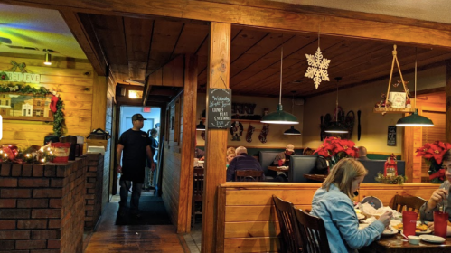 A cozy restaurant interior with wooden decor, diners at tables, and a server walking through a doorway. Holiday decorations visible.