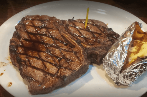 A grilled steak with a crisscross pattern, served on a white plate alongside a foil-wrapped baked potato.