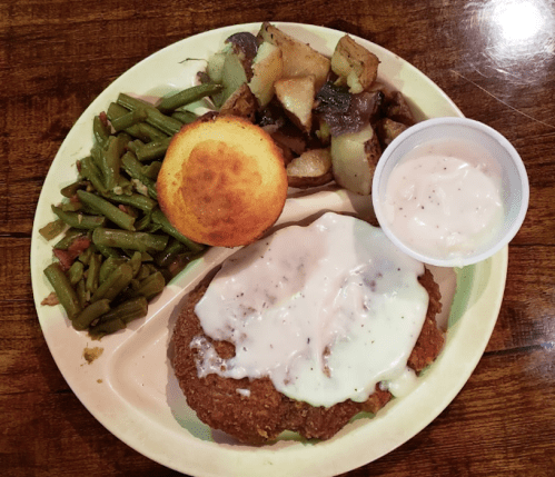 A plate of food featuring fried chicken with gravy, green beans, cornbread muffin, and seasoned potatoes.