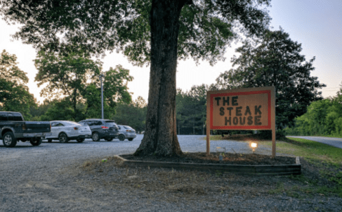 A sign for "The Steak House" next to a gravel parking lot with several cars and trees in the background at dusk.