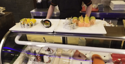 Two plates of sushi on a counter, featuring various rolls and a hand preparing sushi in the background.