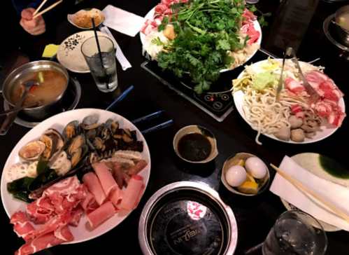 A table set with various dishes for hot pot, including meats, seafood, vegetables, and dipping sauces.