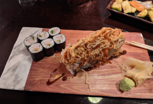 A wooden platter with sushi rolls, a noodle sushi roll topped with sauce, pickled ginger, and wasabi.