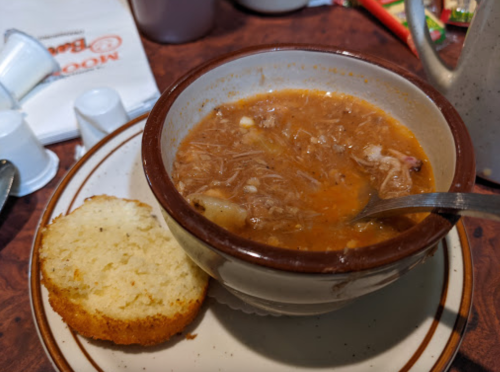 A bowl of hearty soup with shredded meat, served with a slice of cornbread on the side.