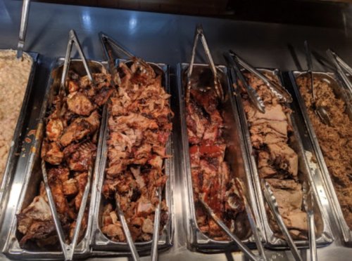 A variety of grilled meats displayed in metal trays with tongs, showcasing different cuts and preparations.