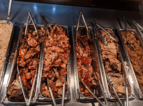 A variety of cooked meats displayed in metal trays with tongs, showcasing different textures and colors.