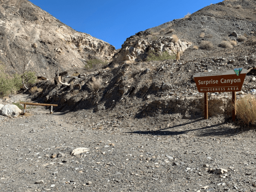 Sign for Surprise Canyon Wilderness Area with rocky terrain and dry vegetation under a clear blue sky.