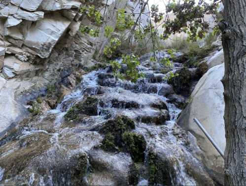 A cascading waterfall flows over rocky steps, surrounded by greenery and rugged cliffs.