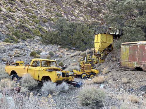 Abandoned yellow truck and machinery in a rugged, overgrown landscape with rocky terrain and sparse vegetation.