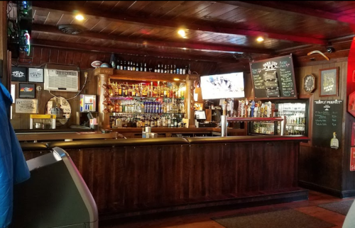 A cozy bar interior featuring a wooden counter, shelves of liquor, and a TV, with warm lighting and a welcoming atmosphere.