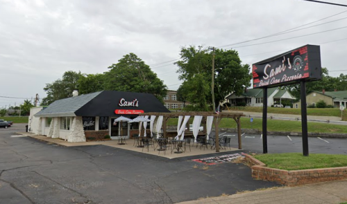 Exterior view of Sam's pizza restaurant with outdoor seating and a sign, set in a suburban area on a cloudy day.