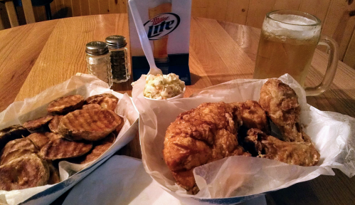 A table with fried chicken, grilled potatoes, coleslaw, and a beer in a glass, set in a cozy restaurant.