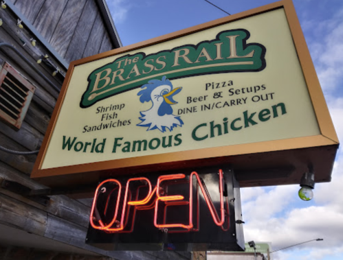Sign for "The Brass Rail" featuring shrimp, fish, sandwiches, pizza, and "World Famous Chicken," with an "OPEN" neon light.