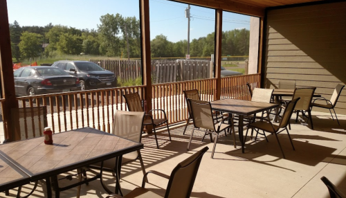 A sunny outdoor patio with empty tables and chairs, surrounded by greenery and a parked car in the background.