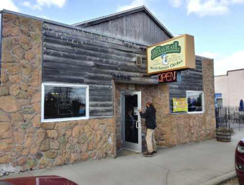 A person stands outside a rustic stone and wood restaurant with an "Open" sign, labeled "Brass Rail - World Famous Chicken."