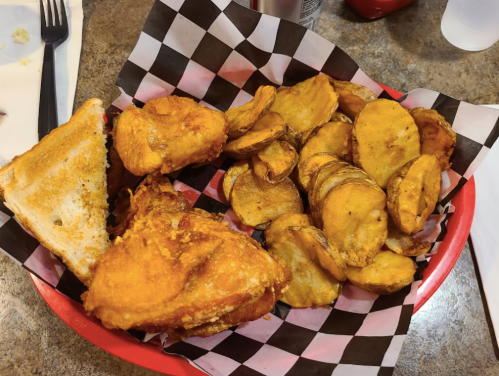 A red plate with fried chicken, golden potato wedges, and a slice of toasted bread on a checkered paper liner.